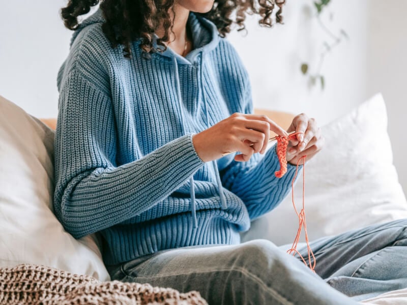 A woman wearing a blue jumper is sitting on a sofa crocheting
